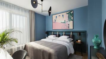 A modern bedroom with a large bed, a side table, a chair, a potted plant, and a wall-mounted artwork.