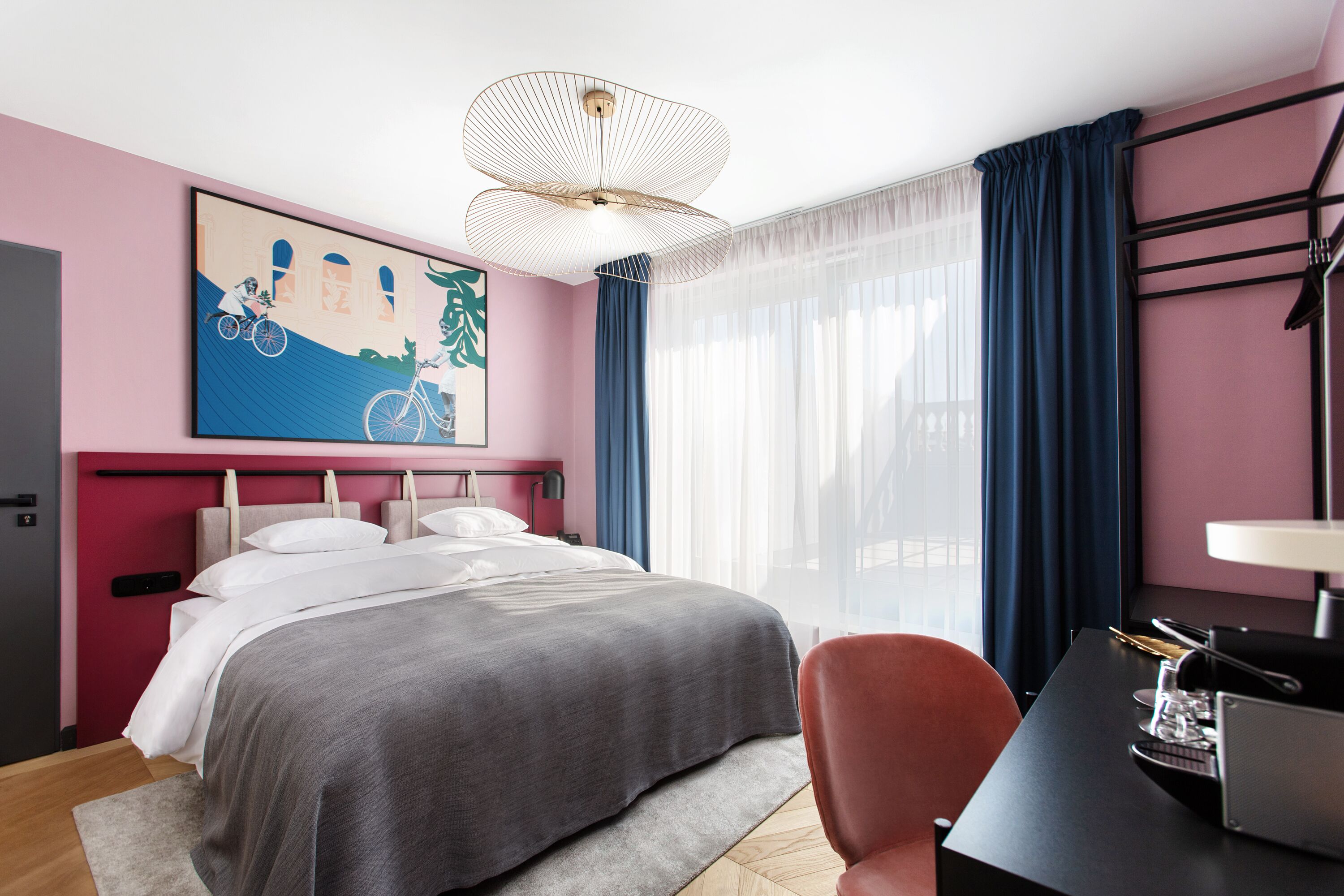 Una habitación de hotel moderna con cama, escritorio, silla, un amplio ventanal con cortinas y una lámpara de techo original.