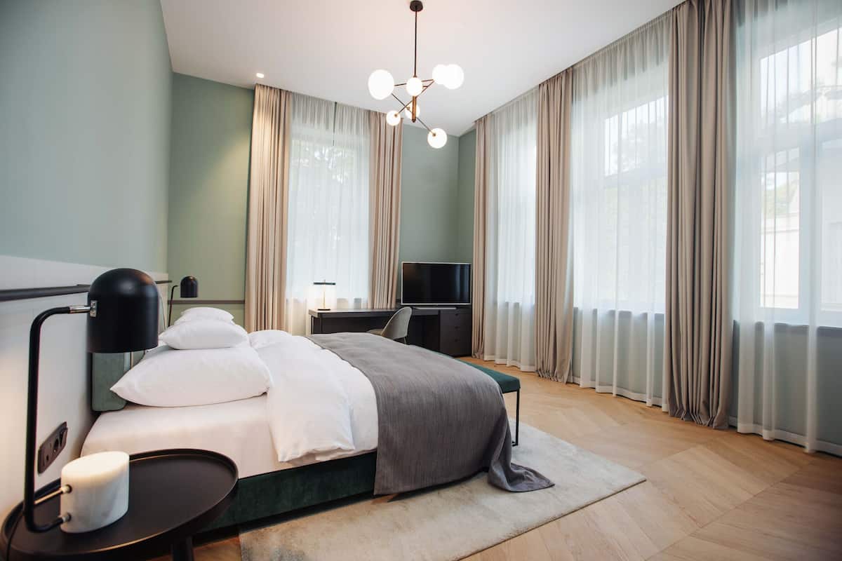 Una habitación de hotel moderna con cama, mesita de noche, lámpara, televisor y cortinas.
