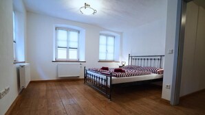 Een slaapkamer met houten vloer, een bed met gestreept beddengoed, twee ramen en een radiator.