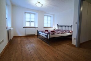 Une chambre avec un plancher en bois, un lit avec une literie à rayures, deux fenêtres et un radiateur.
