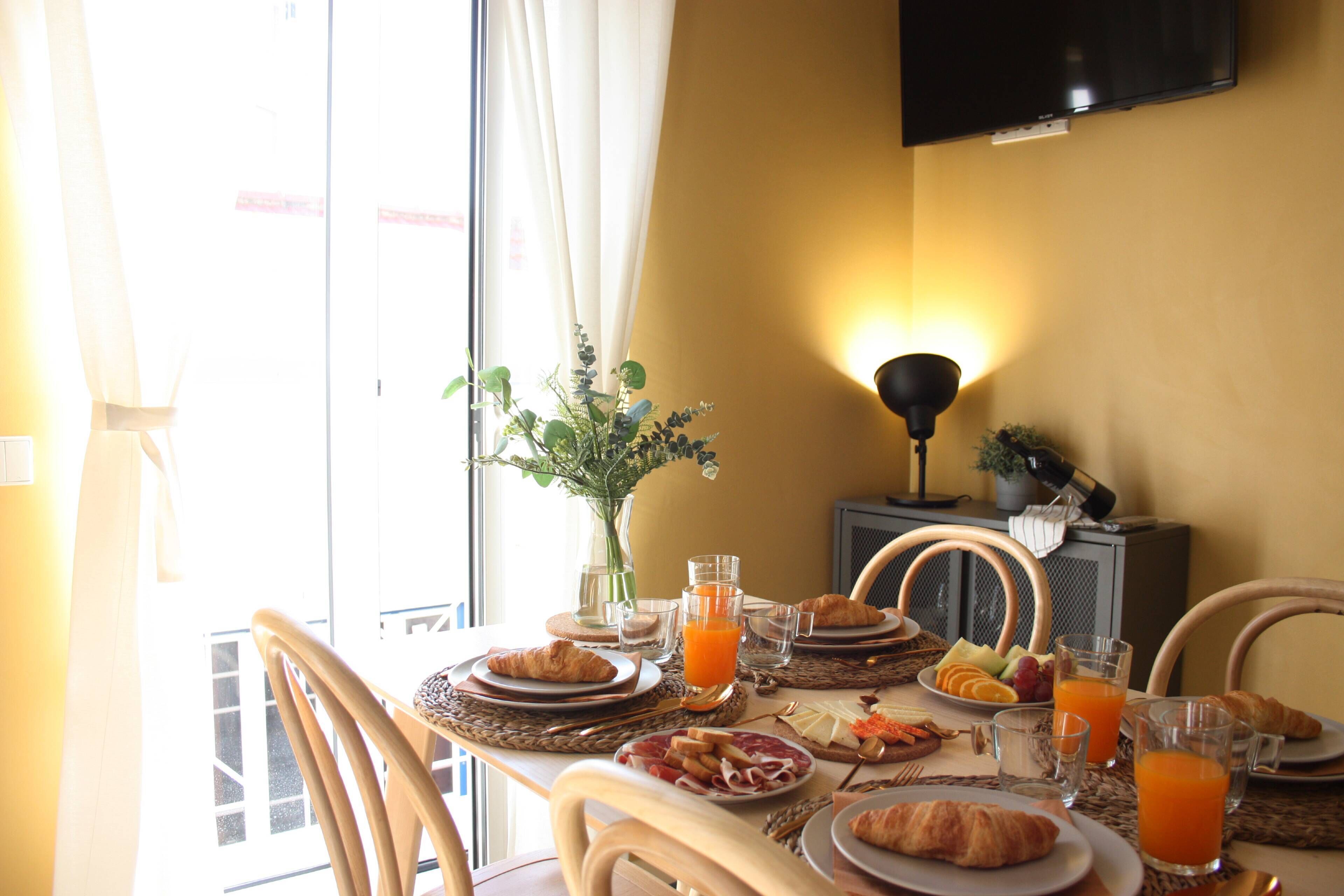 A breakfast table with croissants, fruit, and juice, a vase with greenery, and a TV mounted on the wall.