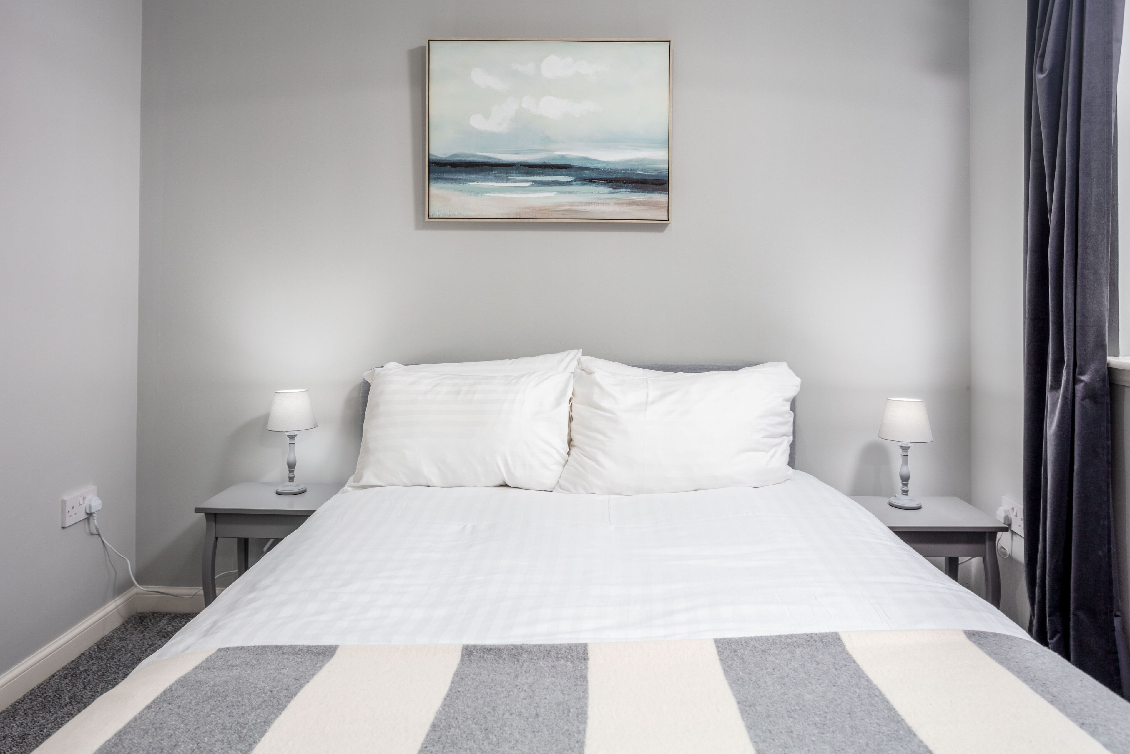 A neatly made bed with white bedding and a striped rug, two bedside tables with lamps, and a painting of a landscape on the wall.
