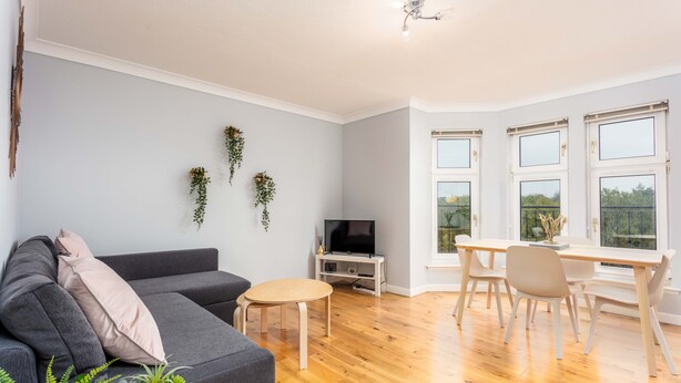 A modern living room with a grey sofa, a small round table, a TV, and a dining area with a white table and chairs.