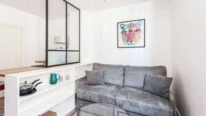 A modern living space with a grey sofa, a white shelving unit with kitchenware, a large mirror, and a colorful abstract painting on the wall. - Newly Renovated, Modern Studio Flat in Montmartre (Paris)