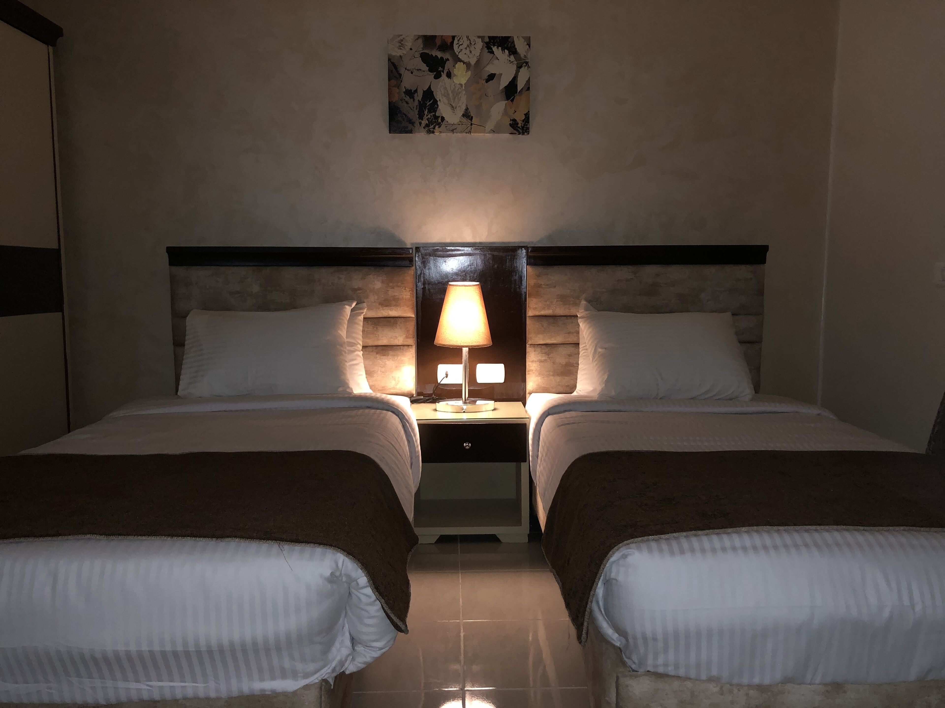 Una habitación de hotel con dos camas, una mesita de noche con lámpara, una obra de arte en la pared y piso de baldosas.