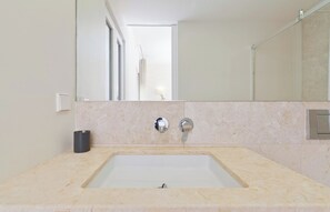 Un baño con un lavamanos rectangular, dos grifos, un espejo y un dispensador de papel higiénico.