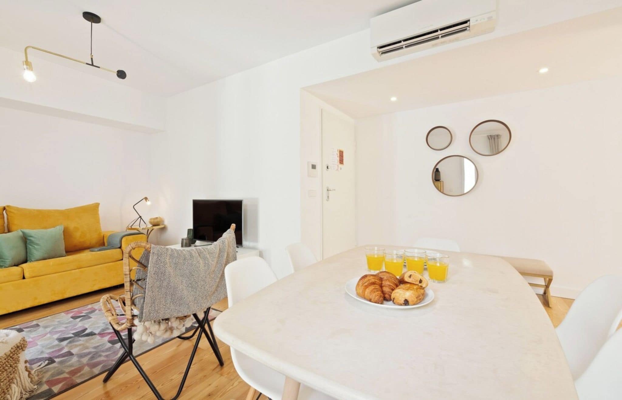 A modern living room with a yellow sofa, a dining table with croissants and orange juice, and a TV.