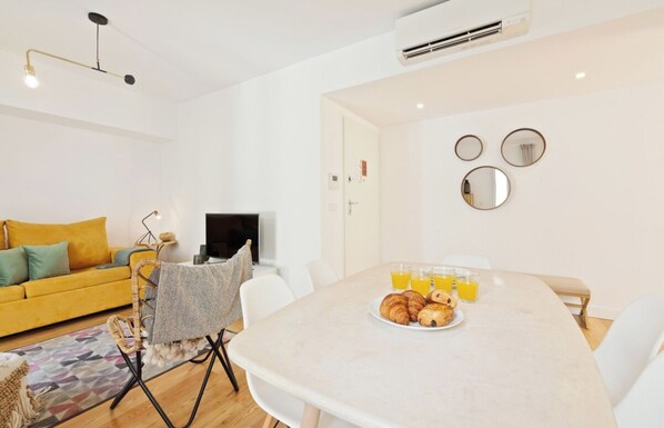 A modern living room with a yellow sofa, a dining table with croissants and orange juice, and a TV.