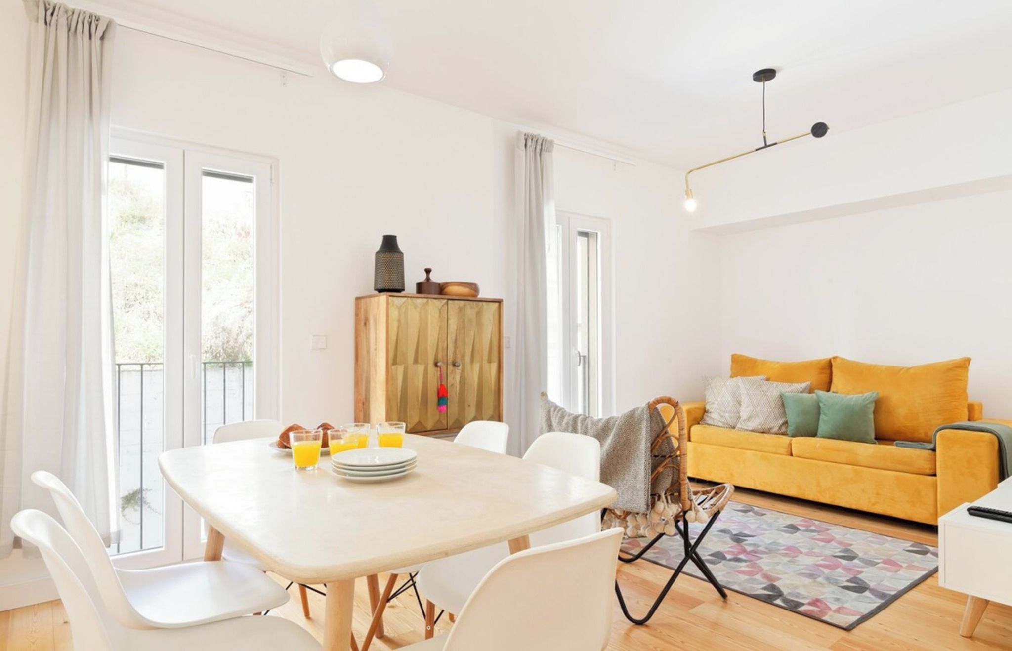 A bright living room with a yellow sofa, a dining table with chairs, and a wooden cabinet.