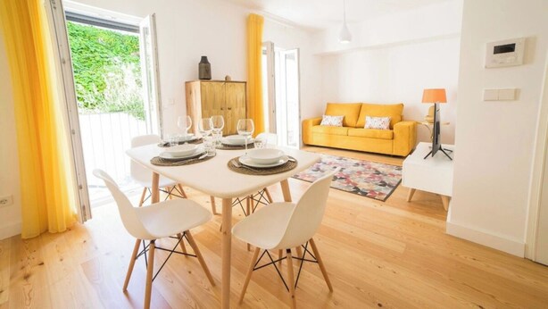 A dining area with a table set for four, a sofa, and a TV stand in a bright room with wooden flooring.