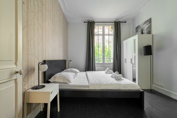 A bedroom with a bed, bedside table, lamp, wardrobe, and a window with curtains. - Vue exceptionnelle sur la Tour Eiffel, ap 3 ch (Paris)