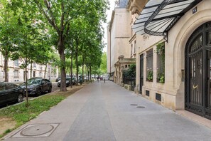 Una acera bordeada de árboles, con coches aparcados y un edificio con amplias ventanas y una verja negra.