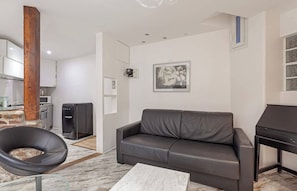 A living room with a grey sofa, a black chair, a marble coffee table, a kitchen area with a microwave, and a wooden beam. - The Modern Eiffel 2BDR Stay in Paris Center (Paris)