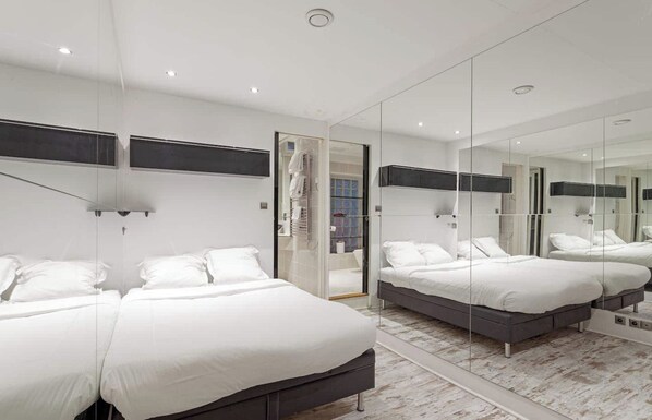 A modern hotel room with a large bed, a separate seating area, a glass partition, and a mirrored wall.