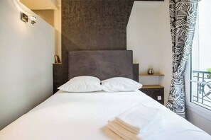 A neatly made bed with white linens, a dark headboard, and a window with patterned curtains. - Beau 2 Pieces Artiste near Saint Germain (Paris)