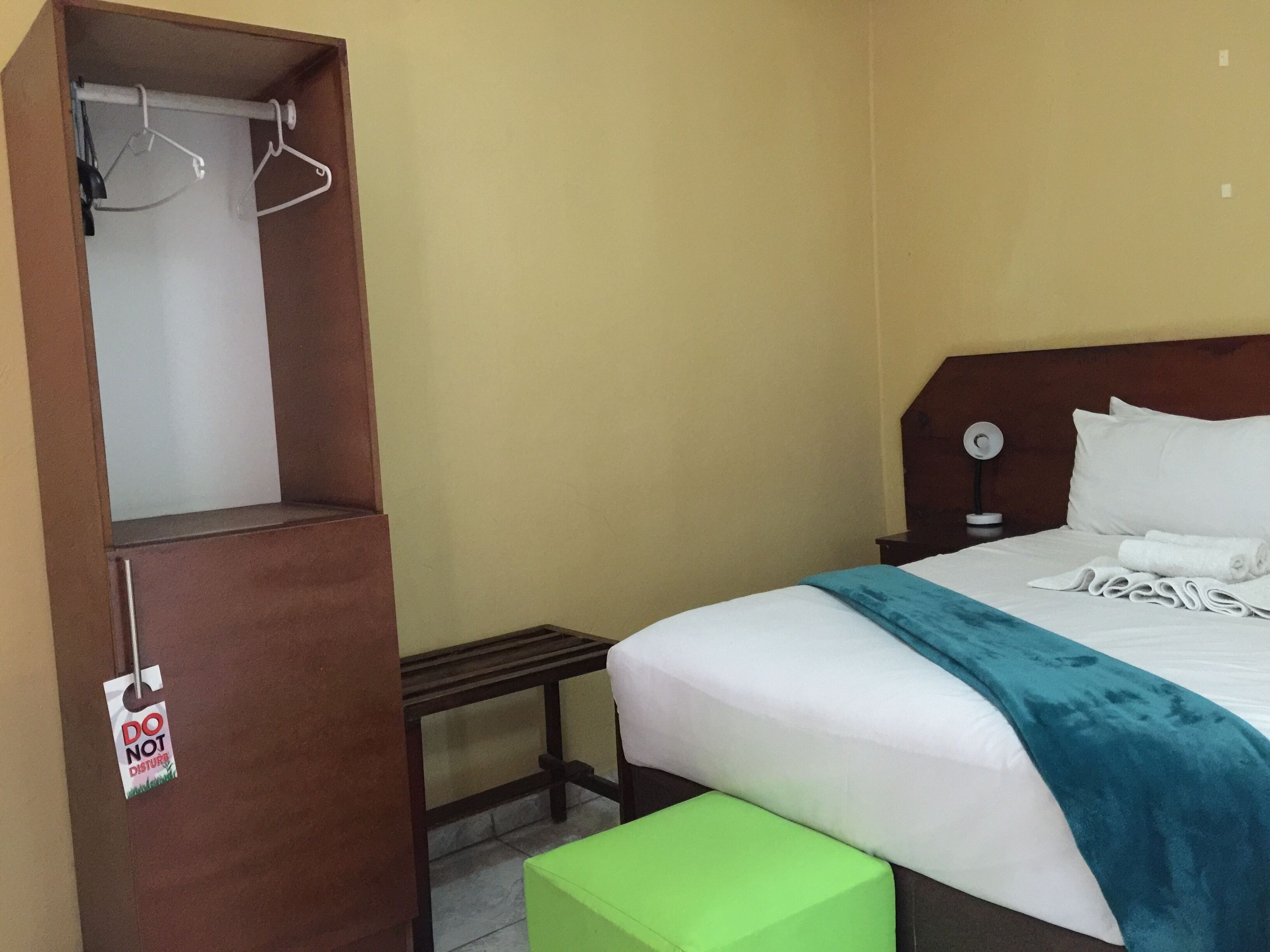 A hotel room with a bed, a green bench, a wardrobe with a 'DO NOT DISTURB' sign, and a bedside table with a lamp.