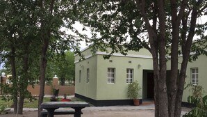 A building with a green exterior and multiple windows, surrounded by trees and a picnic table in a sandy area.