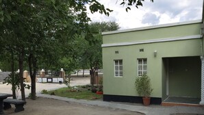A small building with a green exterior and two windows, surrounded by trees and a sandy area.
