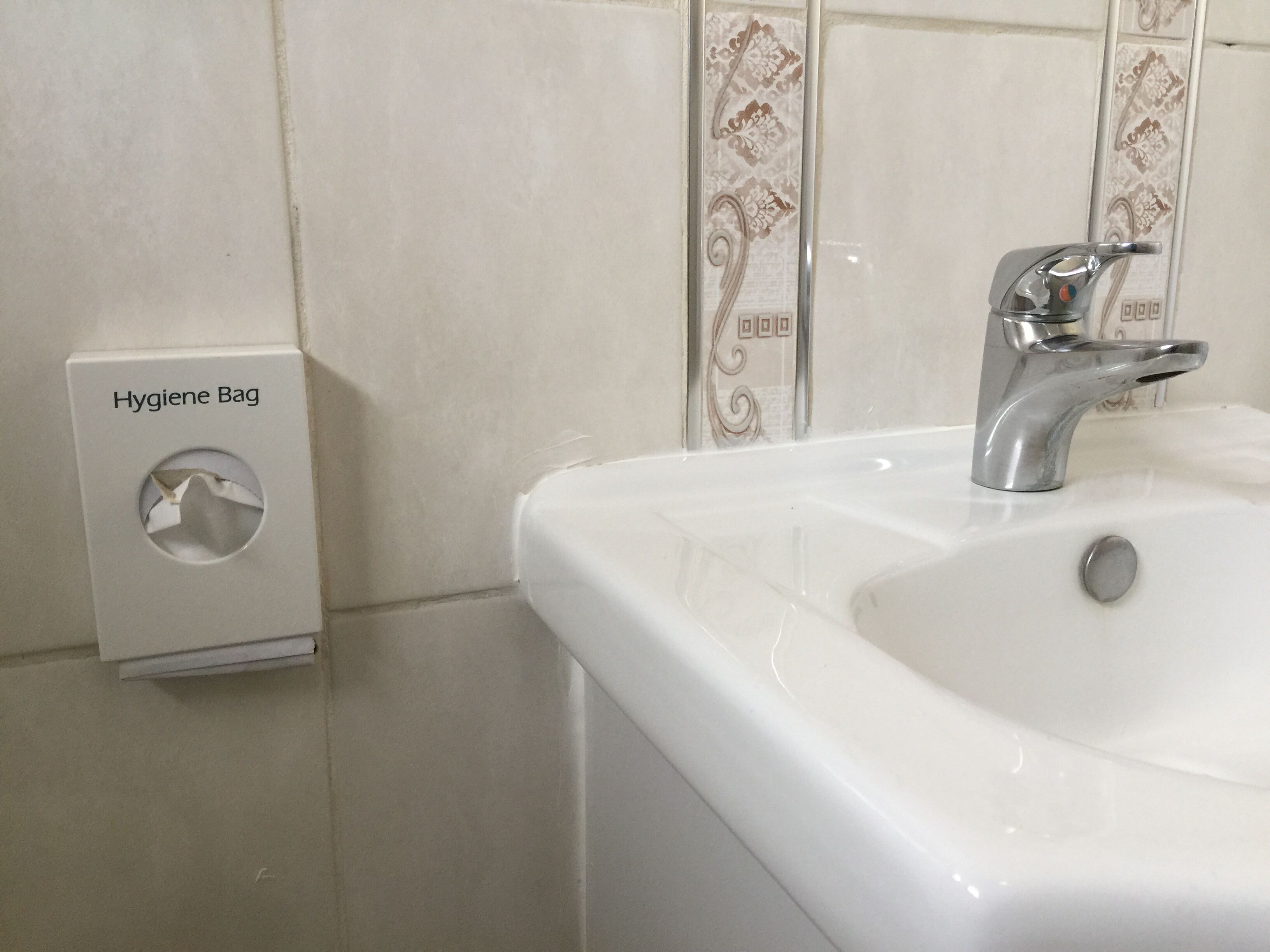 Une salle de bain avec un lavabo blanc, un robinet argenté et un distributeur de sacs d’hygiène fixé au mur.