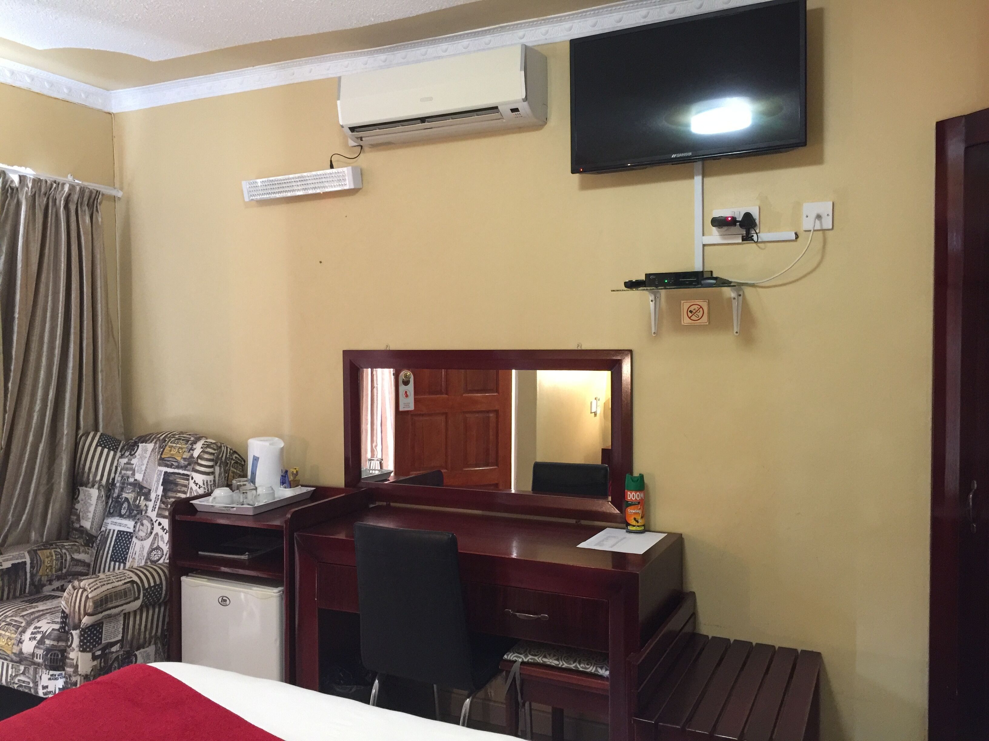A hotel room with a bed, a desk with a chair, a television mounted on the wall, an air conditioning unit, and a door leading to another room.
