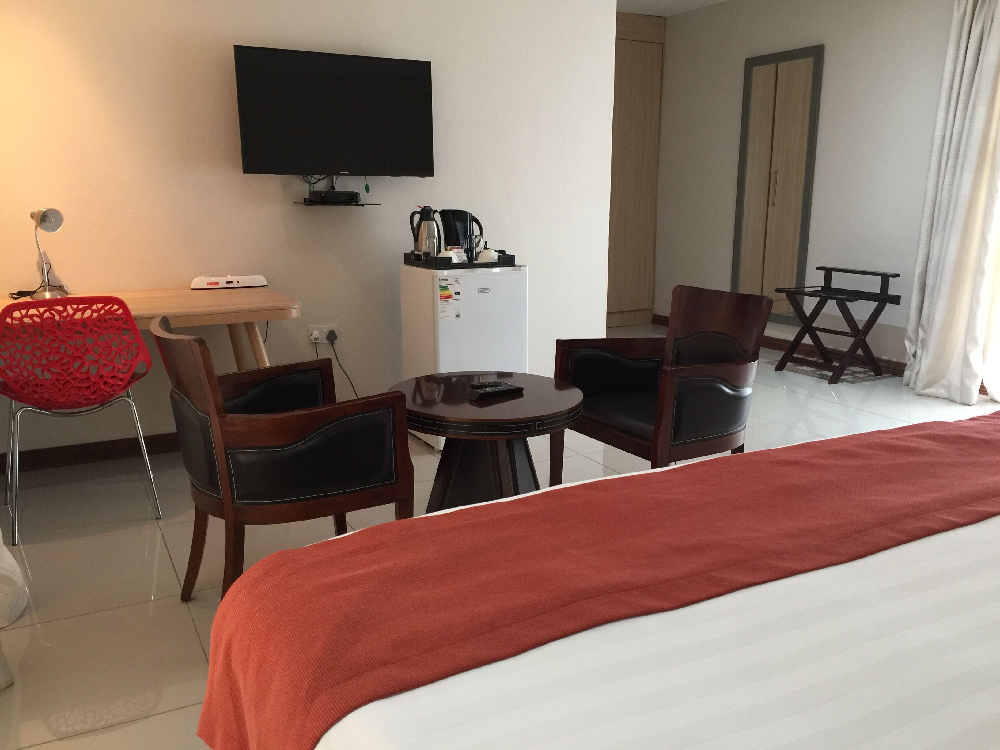Quarto de hotel com cama, duas cadeiras, uma mesinha, televisão, mini-geladeira e uma poltrona vermelha.