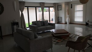 A modern living room with a grey sofa, a matching armchair, and a coffee table. There are large windows with curtains, a potted plant, and a framed picture on the wall. - Royal Court Guest House (Letlhakane)
