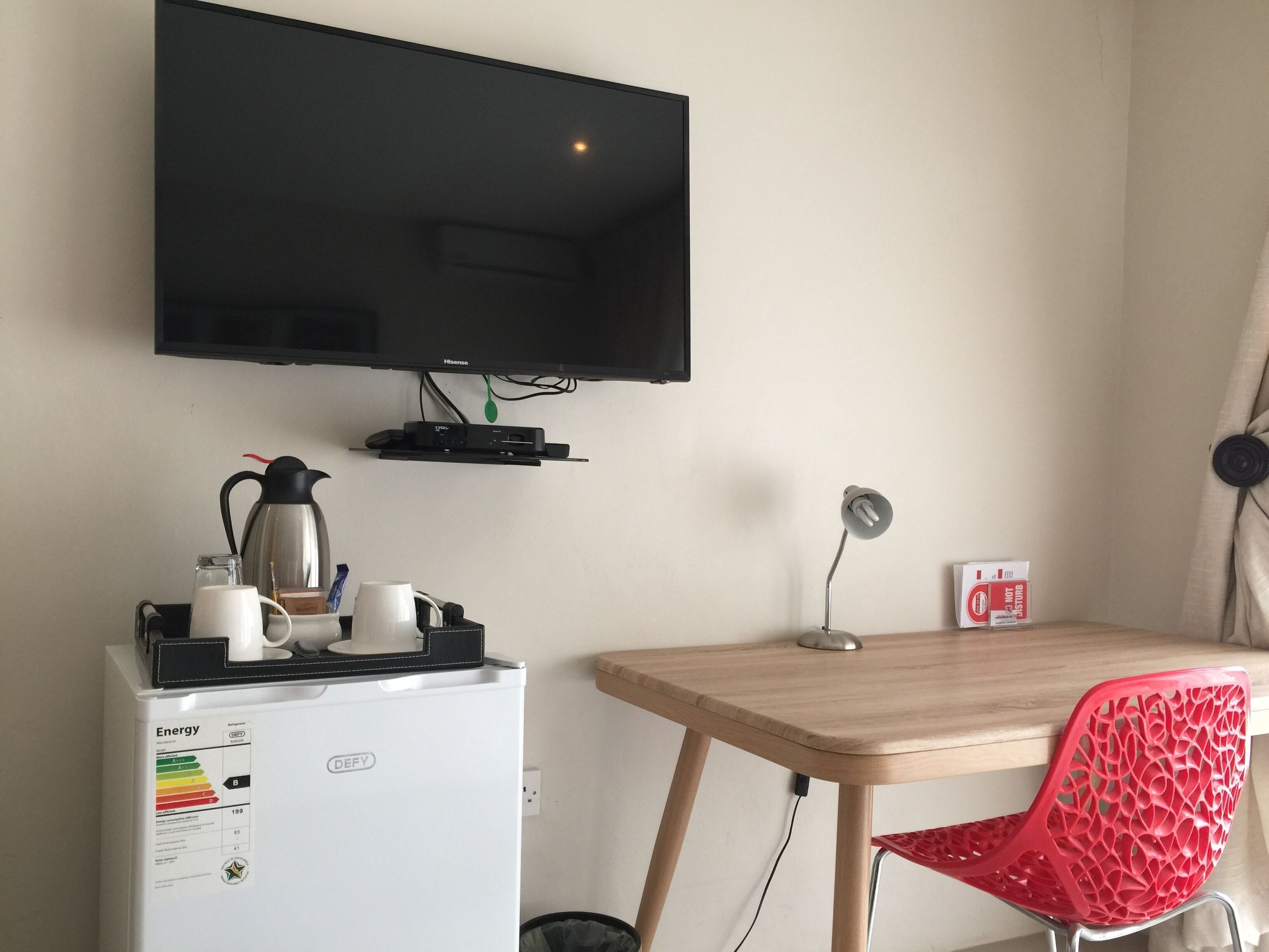 Une chambre compacte dotée d’un téléviseur à écran plat fixé au mur, d’un petit réfrigérateur muni d’étiquettes indiquant son efficacité énergétique, d’un bureau en bois, d’une chaise rouge et d’une petite table avec une lampe.