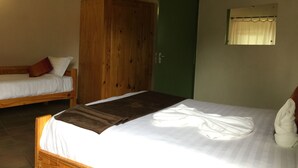 Une chambre avec deux lits, l’un recouvert d’une couverture marron et l’autre d’une couverture blanche. Il y a une porte verte et une fenêtre avec un rideau.