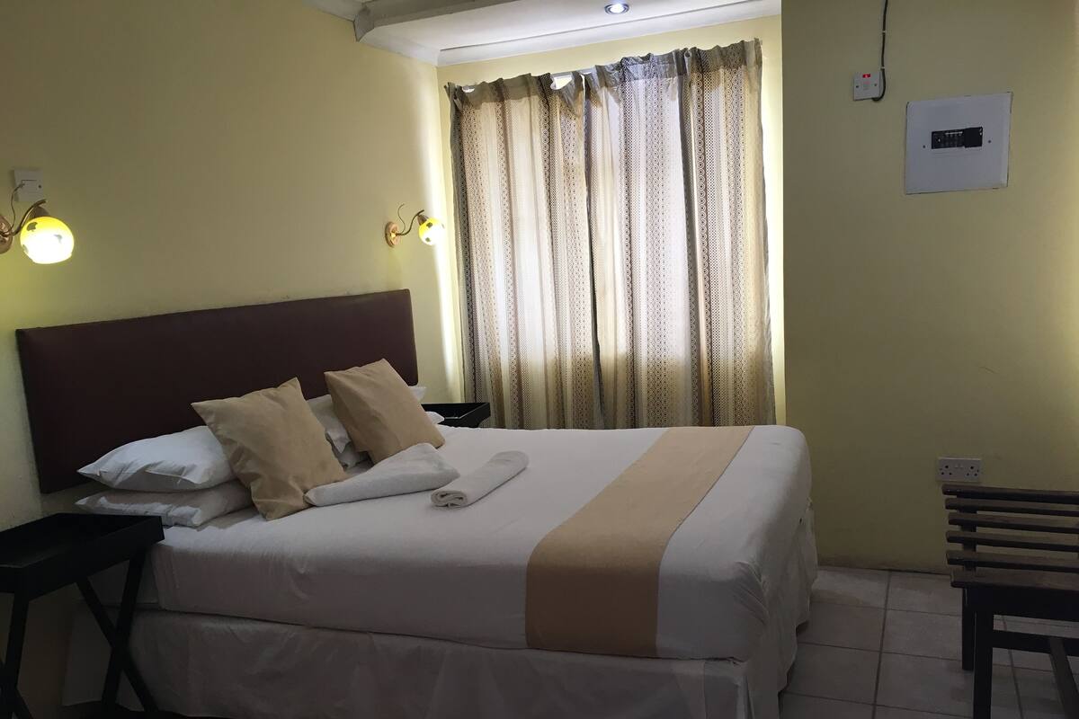 A neatly made bed with a headboard, pillows, and a rolled towel. There is a nightstand with a lamp, a window with curtains, and a wall socket.