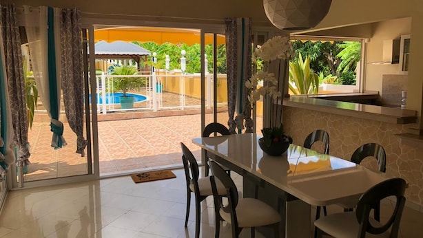 A dining area with a table and chairs, a large window with curtains, and a view of a pool and greenery outside.