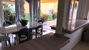A dining area with a long table and chairs, a potted plant, and a view of a garden and building outside.