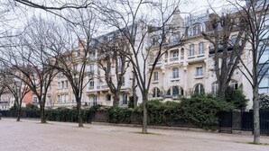 A row of multi-story buildings with balconies, a paved area in front, and trees without leaves.