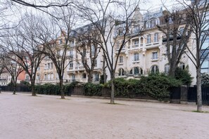 A row of multi-story buildings with balconies, a paved area in front, and trees without leaves.