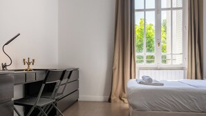 A bedroom with a bed, a desk with a lamp, a chair, and a window with curtains.