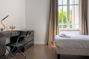 A bedroom with a bed, a desk with a lamp, a chair, and a window with curtains.