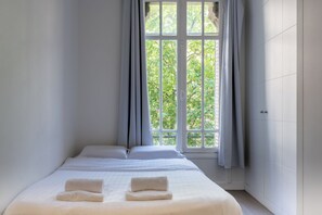 A single bed with white linens and two towels, a window with curtains, and a view of greenery outside. - Gorgeous 2BD flat with view on Champ de Mars! (Paris)