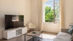 A modern living room with a flat-screen TV on a white cabinet, a glass coffee table, a white chair, and a large window with a view of trees.