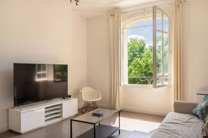 A modern living room with a flat-screen TV on a white cabinet, a glass coffee table, a white chair, and a large window with a view of trees.