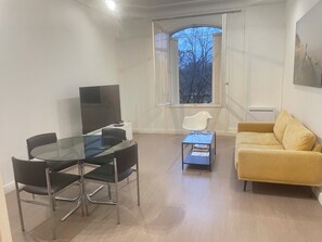 Un salon moderne avec une table à manger en verre, des chaises noires, un canapé jaune, une table basse et un téléviseur.
