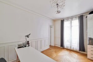 A room with a white table, a lamp, a wardrobe, and a large window with curtains. - Cosy flat for 2 people near Pigalle (Paris)