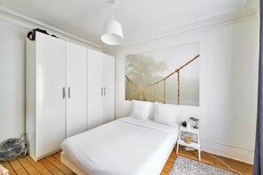 A bedroom with a bed, white wardrobe, a hanging light fixture, a wall-mounted painting of a bridge, and a small bedside table with a lamp.