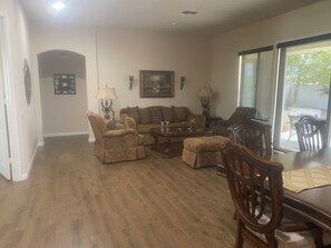 A living room with a sofa, armchairs, a coffee table, and a dining table with chairs. There is a painting on the wall and a window with a view outside.