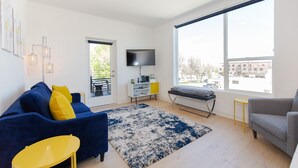 Un salon moderne avec un canapé bleu, un fauteuil jaune, un fauteuil à oreilles gris, une grande fenêtre et un téléviseur à écran plat.