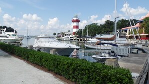 A marina with various boats docked, a lighthouse in the background, and a well-maintained hedge along the walkway.
