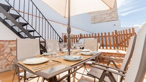 Un área de comedor al aire libre con una mesa preparada para cuatro personas, rodeada de sillas y una sombrilla que proporciona sombra.