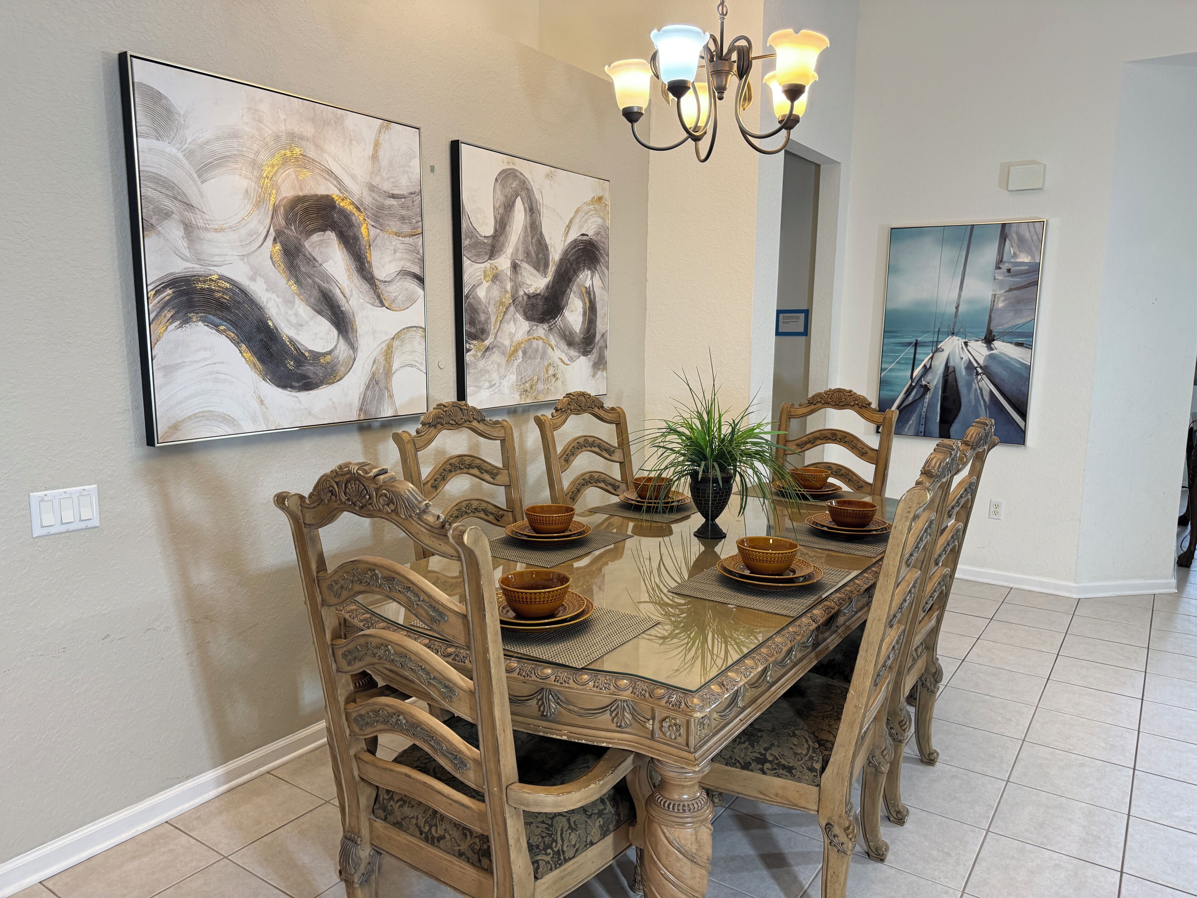 A dining room with a glass-top table, ornate wooden chairs, and artwork on the walls.