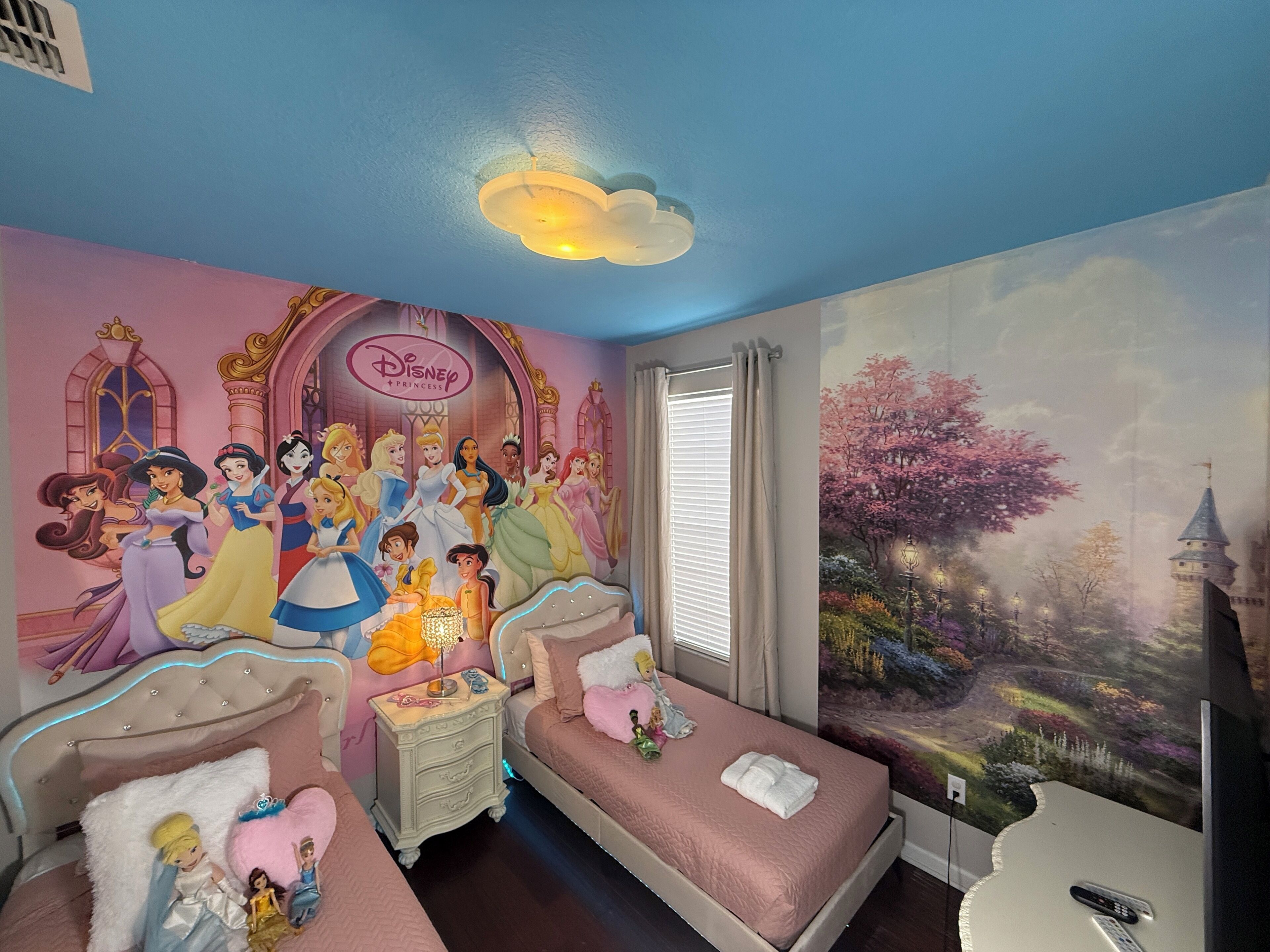 A bedroom with a Disney Princess mural, two beds, a nightstand, and a large window with curtains.