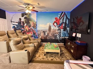 Une salle de cinéma maison avec une grande fresque murale, des fauteuils inclinables en cuir, une table basse et un ventilateur de plafond.