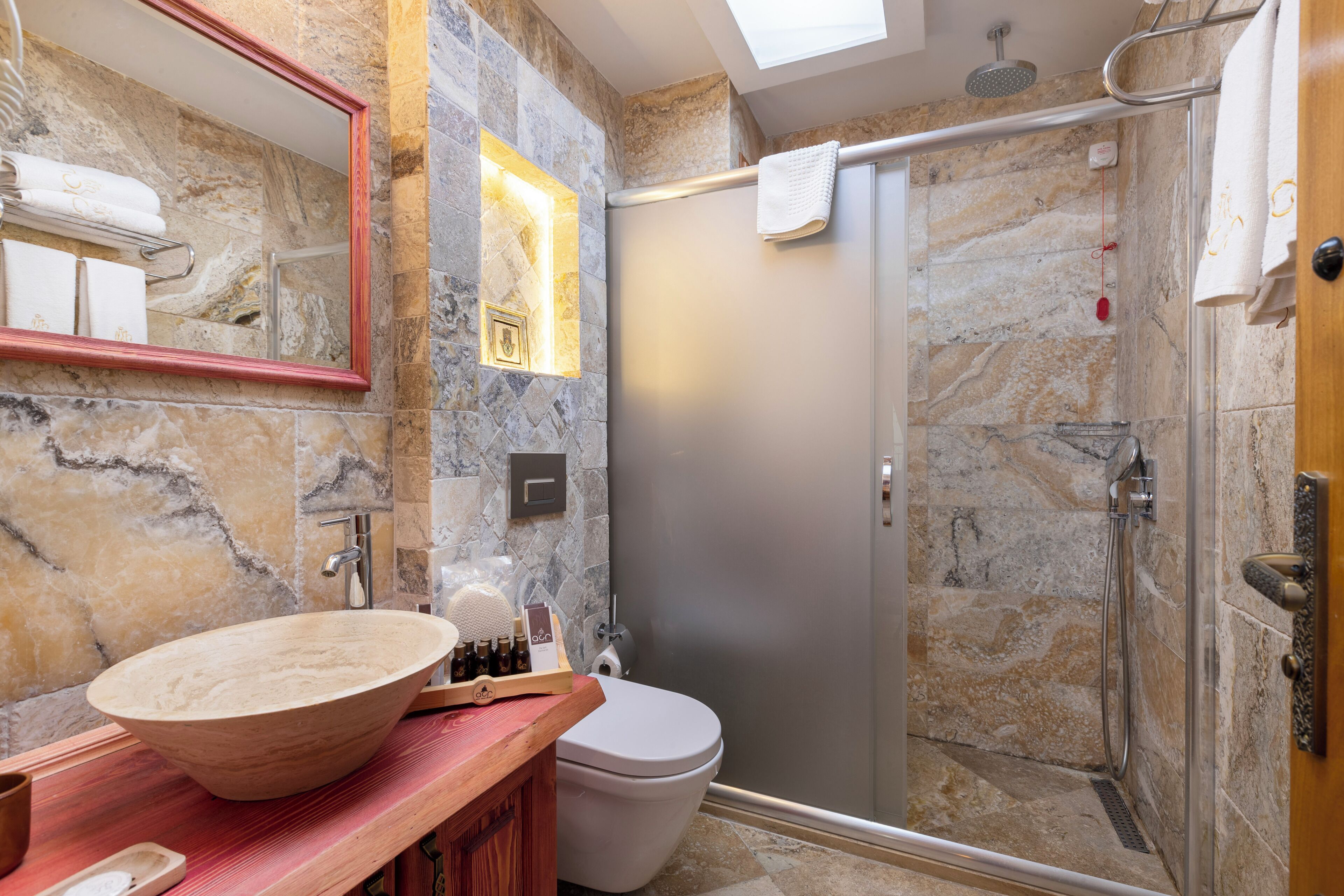 Ein Badezimmer mit Marmorwand, Waschtisch aus Holz, einer großen Schüssel als Waschbecken, Toilette und einer Duschkabine aus Glas.